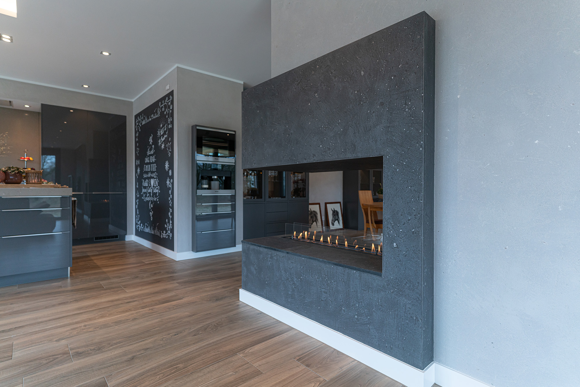 Referenzbild moderner Wohnbereich mit individuell gestalteter Kaminverkleidung in dunklem Frescolori Carmor Putz. Ergänzt durch einem warmen und hochwertigen Holzbodenbelag.