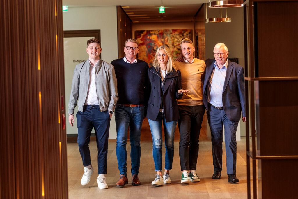 Fünf Personen gehen nebeneinander durch einen modern gestalteten Flur mit warmem Licht und Holzverkleidung an den Wänden. Links läuft ein junger Mann im hellen Jackett und weißen Sneakern, daneben ein Mann mit Brille im dunklen Pullover und Jeans (Chef). In der Mitte geht eine blonde Frau in dunkler Jacke und Jeans (Chefin), rechts daneben ein junger Mann im beigen Pullover und grünen Sneakern, ganz rechts ein älterer Mann im Anzug mit Brille( Senior). Im Hintergrund hängt ein großes buntes Kunstwerk, oben sind grüne Notausgangsschilder zu sehen.