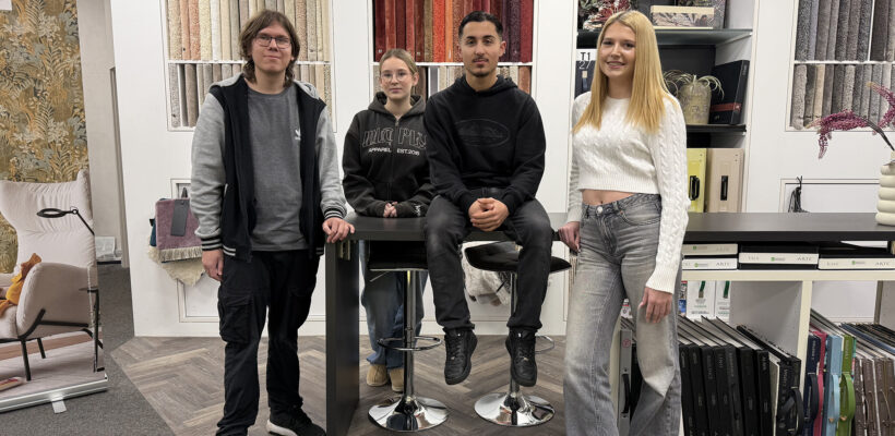 Gruppenfoto der aktuellen Azubis von Ewering im Fachmarkt an einem schwarzen, hohen Beratungstisch. Links steht ein junger Mann mit Brille in schwarzer Jacke und dunkler Hose, in der Mitte steht eine junge Frau mit Brille und dunklem Hoodie hinter dem Tisch. Rechts daneben sitzt ein junger Mann auf dem Tisch, ganz rechts steht eine blonde junge Frau im weißen Strickpullover und hellen Jeans und lehnt am Tisch. Im Hintergrund sieht man eine Wand mit Teppichmustern.