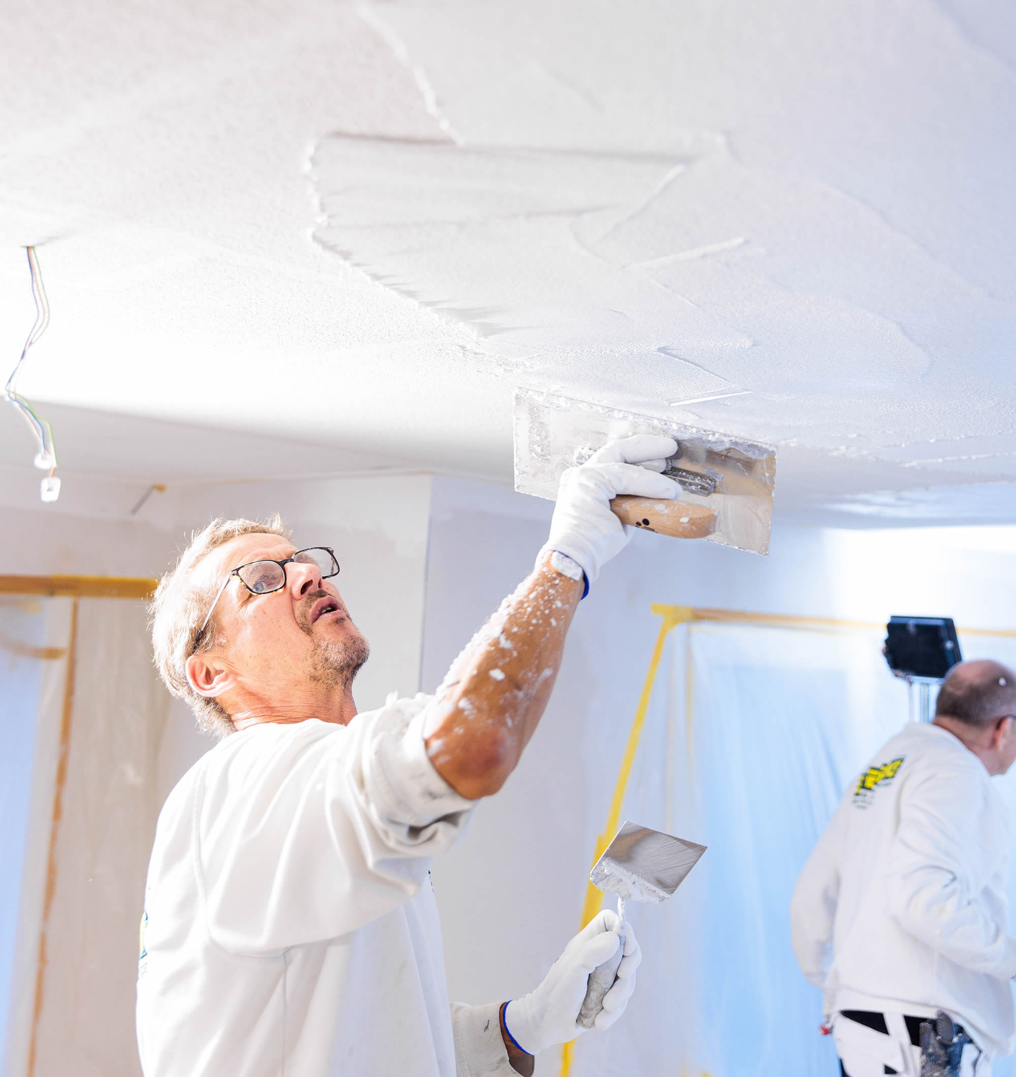 Ewering Mitarbeiter trägt mit einer großen Glättkelle eine weiße Baswa Grundschicht für eine Akustikdecke auf und glättet das Material gleichmäßig. Die Decke zeigt bereits bearbeitete Bereiche mit sichtbaren Kellenzügen, in der anderen Hand hält der Mitarbeiter einen kleineren Spachtel. Im Hintergrund sind Fenster und Wandflächen mit Folie abgeklebt, außerdem steht ein weiterer Mitarbeiter in Arbeitskleidung im Raum und bereitet den nächsten Arbeitsschritt vor.
