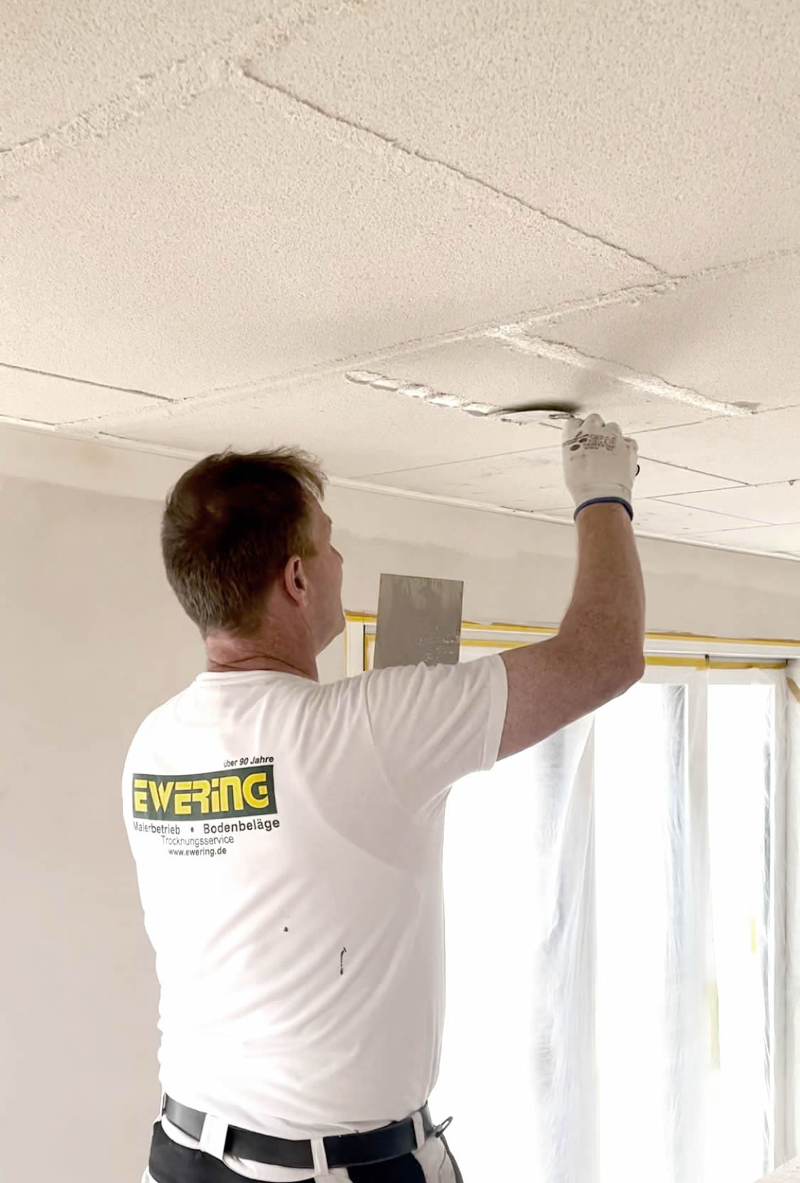 Ewering Mitarbeiter in weißem T Shirt mit Ewering Logo und Arbeitshandschuhen verspachtelt die Fugen von Baswa Akustikplatten an der Decke. Mit einem Spachtel glättet er eine längliche Fugenstelle zwischen den Platten, die Decke zeigt mehrere Plattenfelder mit sichtbaren Stoßkanten. Im Hintergrund sind Fenster mit Folie abgeklebt und die Wände wirken noch im Ausbau.