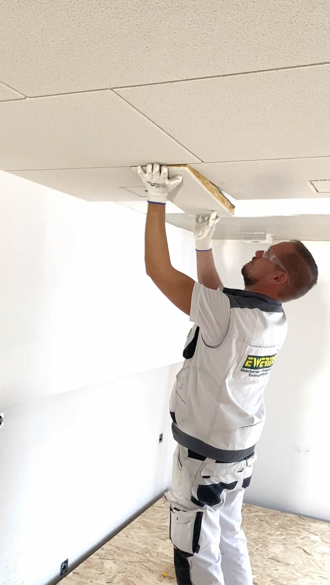 Ewering Mitarbeiter in weißer Arbeitskleidung mit Handschuhen und Schutzbrille montiert Baswa Akustikplatten an der Decke. Er hält eine rechteckige Platte an eine bereits verlegte Deckenfläche mit sichtbaren Plattenfugen und setzt sie passgenau in eine offene Stelle ein. Die Unterseite der Platte wirkt hell und die Kante zeigt das Plattenmaterial, der Raum ist hell und noch im Ausbau mit rohem Boden.
