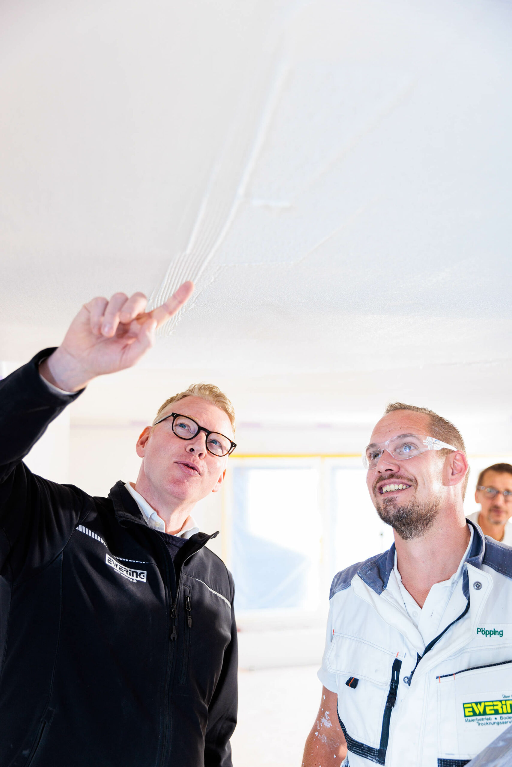 Zwei Personen stehen auf einer Baustelle und schauen nach oben zur Decke, während eine Person mit ausgestrecktem Arm auf eine bearbeitete Fläche zeigt. An der Decke sind helle, gespachtelte Bereiche und streifenförmige Strukturen zu erkennen, die wie vorbereitete Zonen für eine Baswa Akustiklösung wirken. Beide tragen Arbeitskleidung, eine Person hat eine Ewering Jacke an, die andere Schutzbrille und Arbeitsweste,