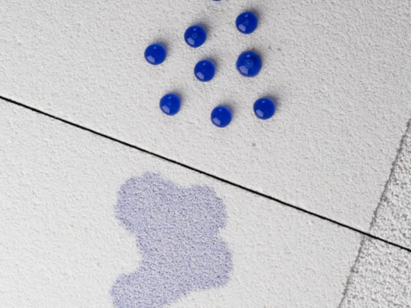 Detailaufnahme einer hellen Baswa Akustikoberfläche mit feiner Struktur und sichtbarer Plattenfuge. Auf der Oberfläche liegen mehrere blaue Tropfen als Wasser oder Schutztest, darunter ist eine bläuliche Verfärbung zu sehen. Das Motiv zeigt Baswa Protect als Oberflächenschutz für Akustiklösungen gegen Feuchtigkeit und Verschmutzung.