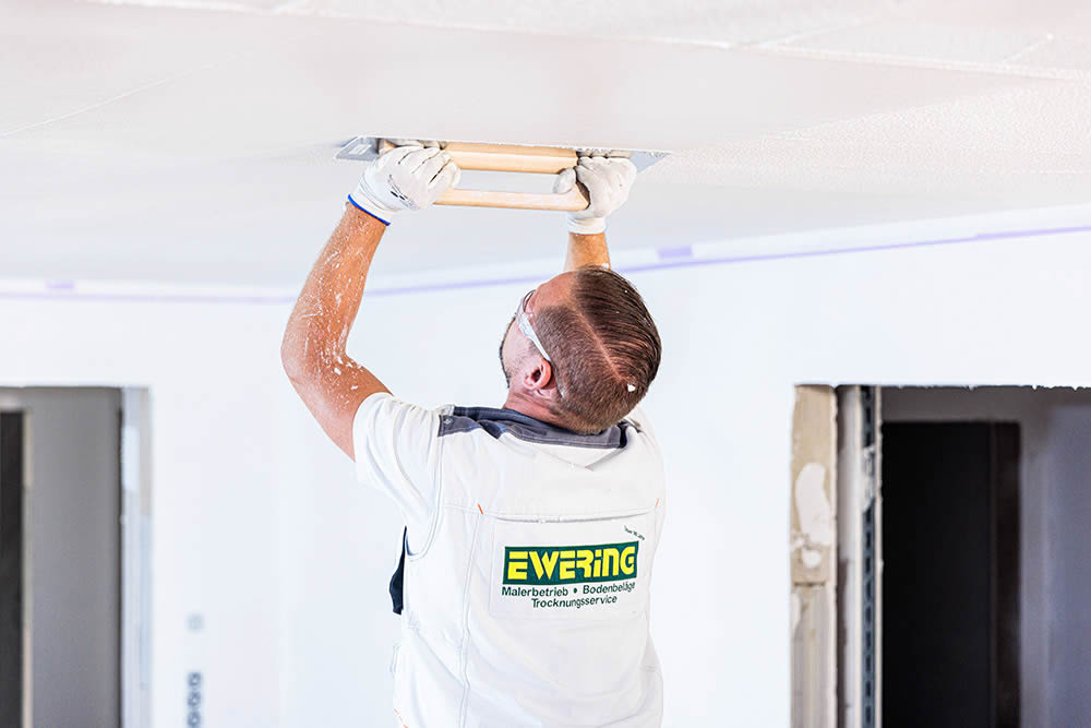 Ewering Mitarbeiter in weißer Arbeitskleidung mit Schutzbrille glättet eine weiße Baswa Akustikbeschichtung an der Decke mit einer breiten Glättkelle. Er arbeitet über Kopf und zieht das Material gleichmäßig ab, die Deckenfläche wirkt bereits fein strukturiert und frisch bearbeitet. Im Hintergrund sind helle Wände, abgeklebt Kanten und offene Türdurchgänge einer Baustelle zu sehen.