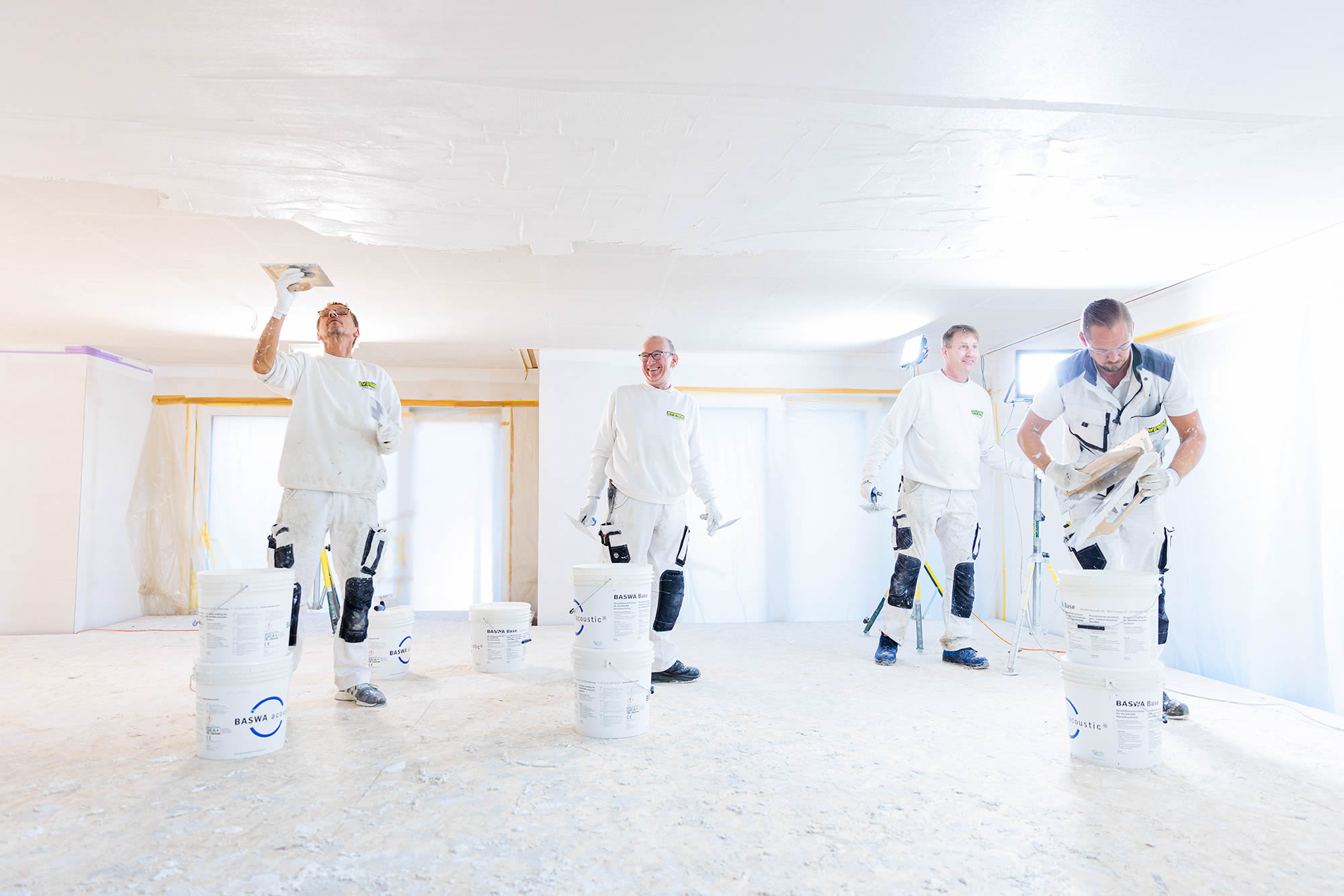 Vier Ewering Mitarbeiter in weißer Arbeitskleidung arbeiten in einem hellen Baustellenraum an einer Decke mit Baswa Akustikbeschichtung. Links glättet ein Mitarbeiter die Deckenfläche mit einer Kelle, in der Mitte stehen zwei Kollegen mit Werkzeug in der Hand, rechts trägt ein Mitarbeiter Material auf einer Traufel oder Glättkelle. Auf dem Boden stehen mehrere weiße Eimer mit Baswa Beschriftung, im Hintergrund sind Fenster und Wandflächen mit Folie und Klebeband abgeklebt und eine Baustellenleuchte sorgt für zusätzliches Licht.