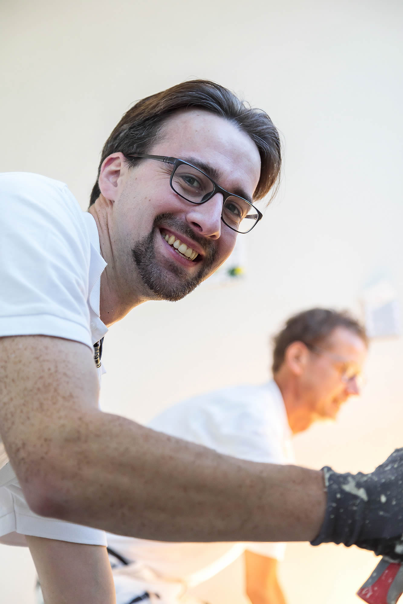Nahaufnahme eines lächelnden Ewering Mitarbeiters mit Brille und kurzem Bart in weißer Arbeitskleidung, der auf einer Baustelle arbeitet. Im Vordergrund ist sein Arm mit Arbeitshandschuh zu sehen, er hält ein Werkzeug in der Hand und schaut direkt in die Kamera. Im Hintergrund arbeitet ein weiterer Mitarbeiter unscharf, die Szene wirkt hell und zeigt Malerarbeiten im Innenbereich.