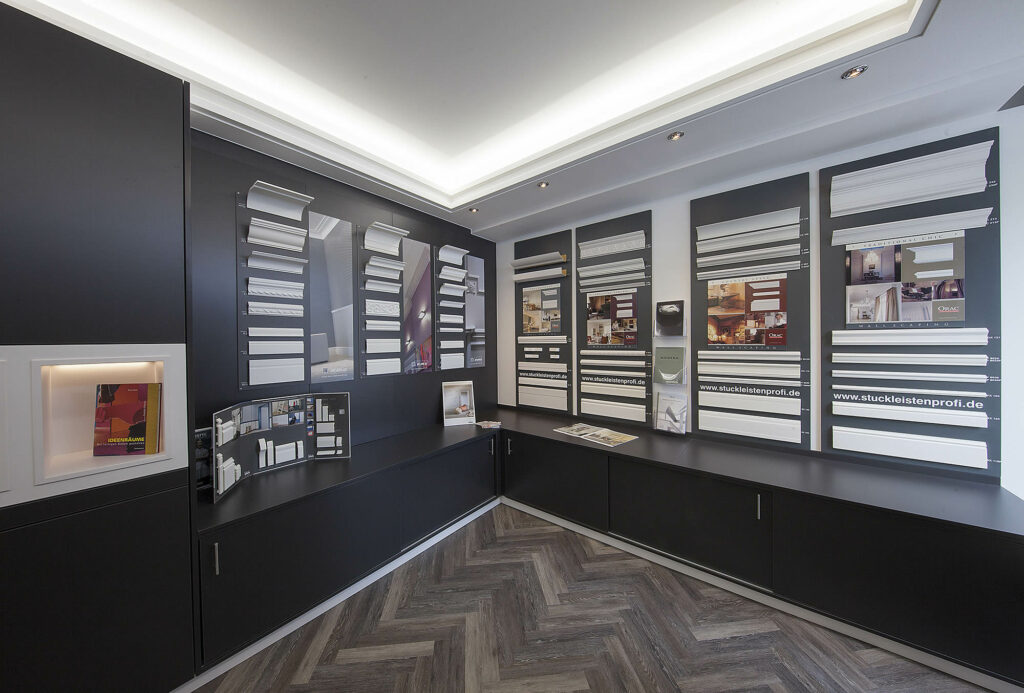 Stuckleisten Ausstellung im Fachmarkt bei Ewering. An den Wänden hängen mehrere große Musterplatten, auf denen viele weiße Stuckleistenprofile in unterschiedlichen Breiten, Formen und Stufen montiert sind, teils mit kleinen Beschriftungen daneben. Auf den Musterplatten sind außerdem Produktbilder und Infotafeln zu sehen, auf der Theke liegen Broschüren und Musterunterlagen.