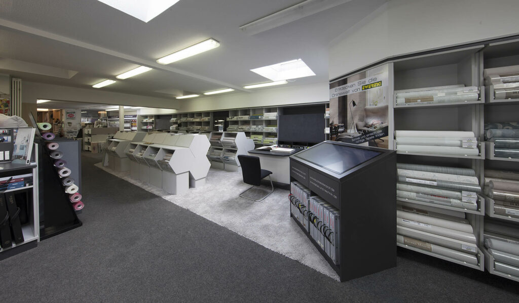 Große Tapetenabteilung im Fachmarkt bei Ewering mit mehreren weißen Tapetenständern in der Mitte des Raums und langen Regalen im Hintergrund. Rechts steht ein hohes Tapetenregal mit vielen Tapetenrollen in verschiedenen Farben und Mustern, davor ein schwarzer Touchscreen Beratungsterminal mit Ordnerablage. Daneben befindet sich ein Beratungstisch mit Stuhl auf einem hellen Teppich, an der Decke sind lange Leuchten und zwei Dachfenster zu sehen.