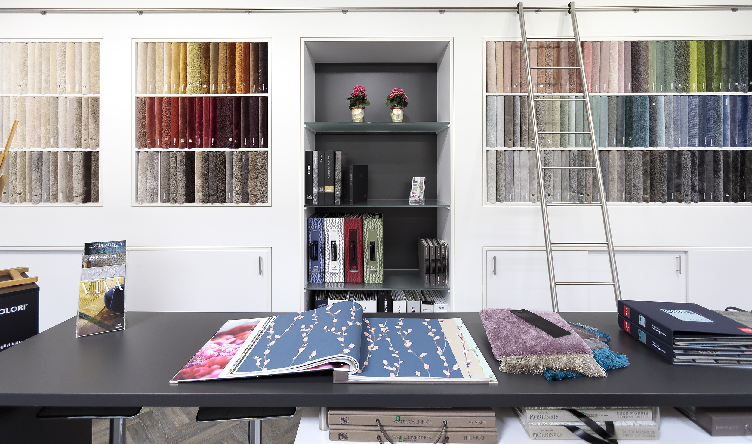 Beratungs- und Ausstellungsbereich im Fachmarkt bei Ewering mit großem Tisch, geöffnetem Tapetenbuch und Stoffmustern. Dahinter eine weiße Musterwand mit Teppichmustern in vielen Farben die farbig sortiert sind.