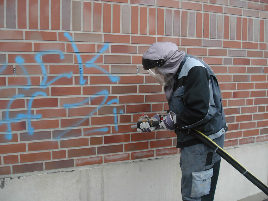 Ein Mitarbeiter in Schutzkleidung mit Haube, Visier und Handschuhen entfernt blaues Graffiti von einer roten Klinkerwand. Er hält eine Strahldüse mit angeschlossenem Schlauch direkt an die Fassade und bearbeitet die besprühten Stellen, unterhalb der Klinker ist ein heller Sockel zu sehen. Das Motiv zeigt die Untergrundreinigung und Graffiti Entfernung an der Außenwand.