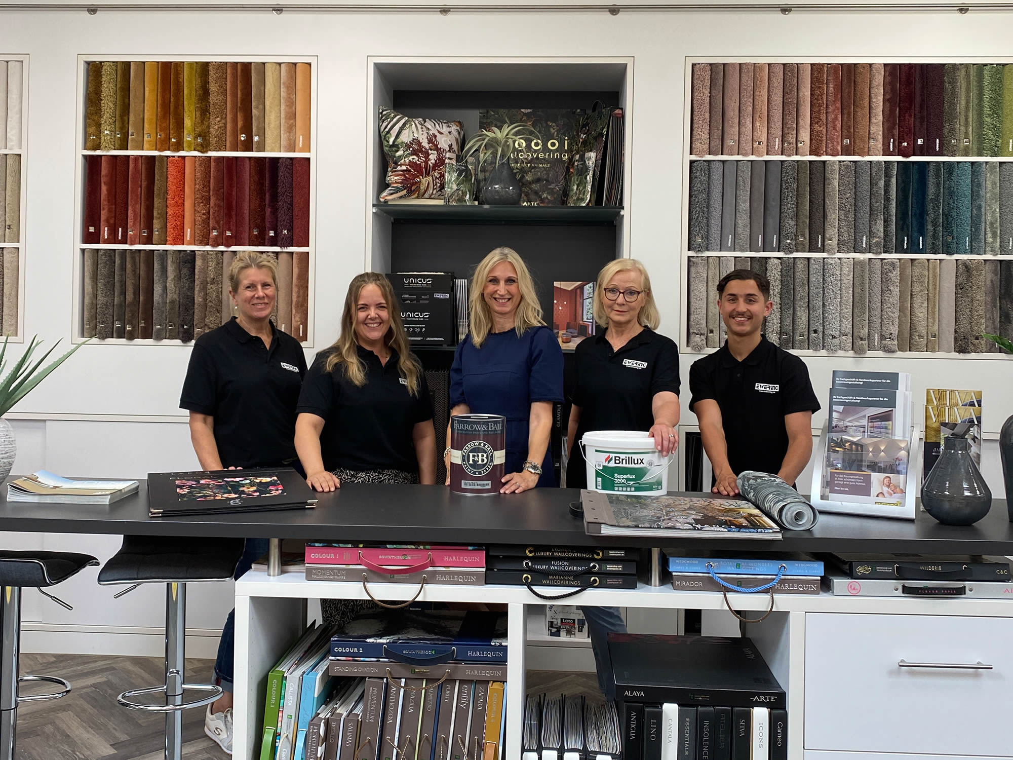 Teamfoto im Ewering Fachmarkt am großen schwarzen Beratungstisch vor farblich sortierten Teppichmusterwänden. Fünf Mitarbeitende stehen hinter dem Tisch, vier tragen schwarze Poloshirts mit Ewering Logo, eine Person trägt ein blaues Oberteil. Auf dem Tisch liegen Tapeten und Materialmuster, außerdem stehen ein Farrow and Ball Farbeimer und ein Brillux Eimer, rechts sind ein Prospektständer, ein gerolltes Muster und eine dunkle Vase zu sehen.
