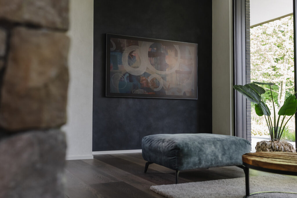 Wohnzimmer mit dunkler dekorativer Frescolori Wand, gerahmtem Bild, gepolstertem Hocker, Pflanze und großen Fenstern.