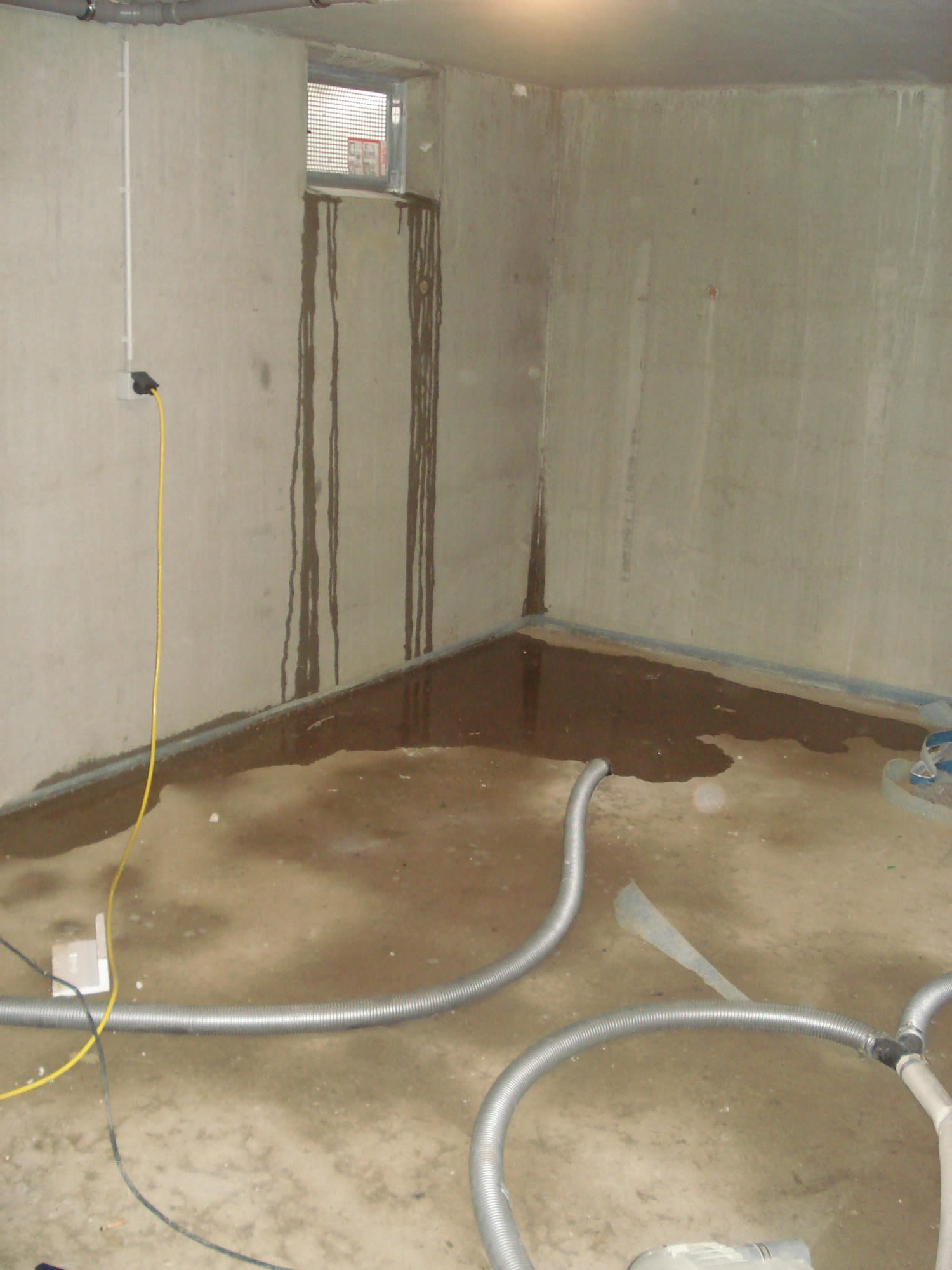 Kellerraum mit feuchten Wänden, Wasser auf dem Boden und Schläuchen für die Trocknung nach einem Wasserschaden.