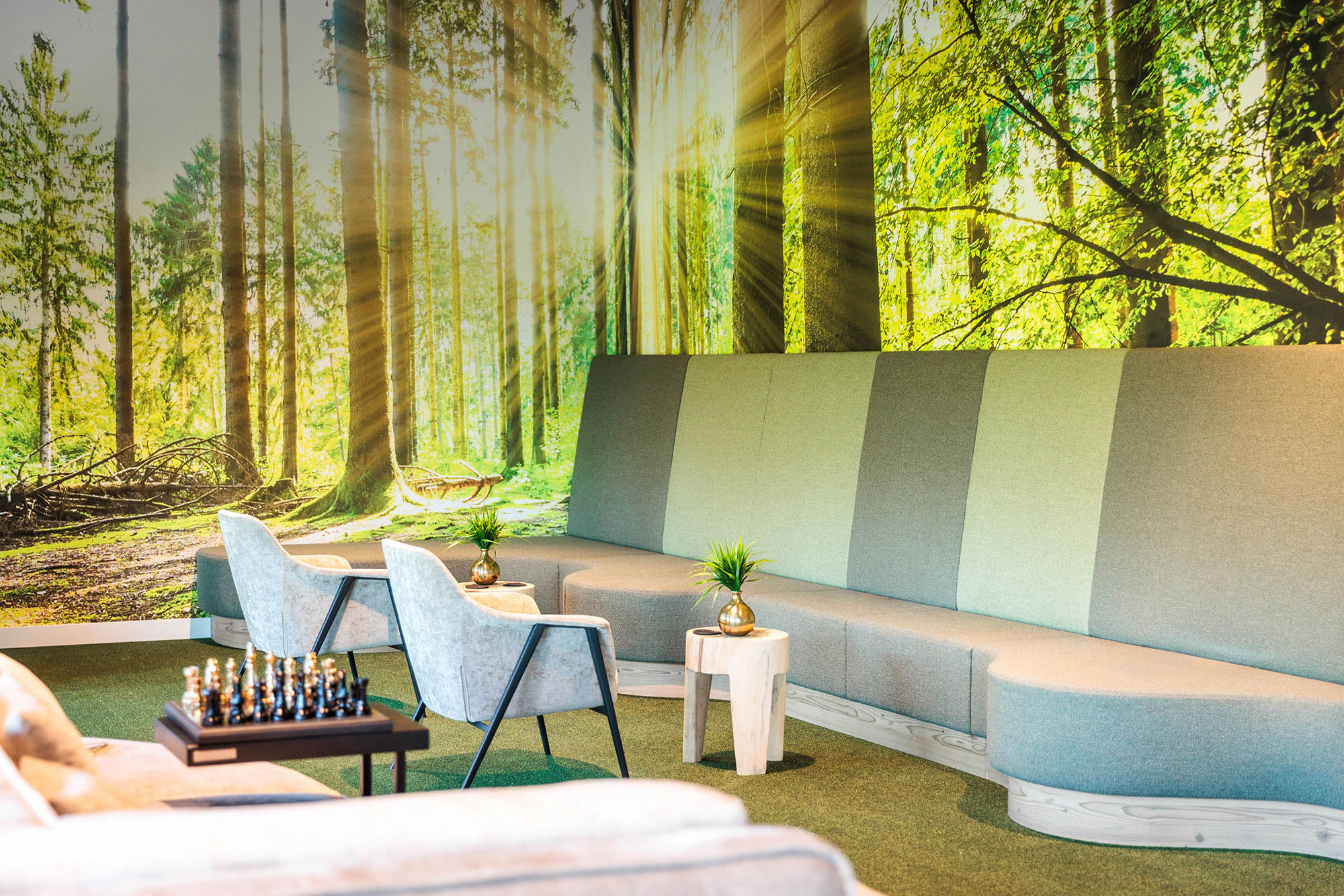 Großflächige Fototapete in einem Wartebereich eines Gewerbeobjekts mit realistischem Waldpanorama. Die Tapete zeigt hohe gerade Baumstämme, grün bewachsenen Waldboden und markante Sonnenstrahlen, die durch den Wald fallen und dem Motiv eine helle, natürliche Tiefenwirkung geben. Im unteren Bereich des Bildes ist ein Teil der Sitzmöbel zu sehen, wodurch erkennbar wird, dass die professionell angebrachte Fototapete den Wartebereich gestalterisch prägt.