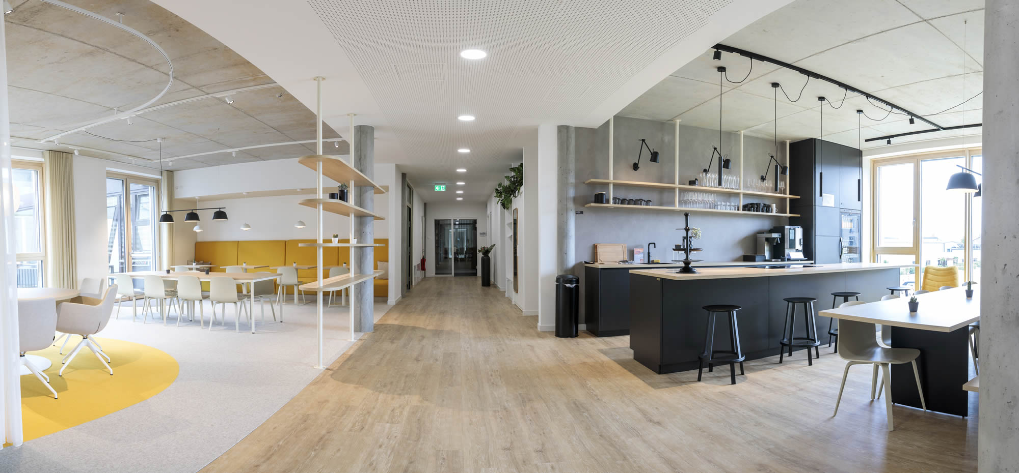 Offen gestalteter Aufenthaltsbereich in einem modernen Gewerbeobjekt mit Büroküche, Essbereich und angrenzender Loungezone. Rechts ist eine große Kücheninsel in dunkler Ausführung mit heller Holzarbeitsplatte, Hockern, offenen Regalböden und Pendelleuchten zu sehen. Links schließen sich helle Sitzplätze, gelbe Akzentflächen, weitere Tische und ein wohnlich gestalteter Aufenthaltsbereich an. Der Raum wirkt durch die Kombination aus heller Möblierung, sichtbarer Betondecke, perforierter Decke im Flurbereich, Holzoptik im Boden und großen Fensterflächen großzügig, modern und einladend.