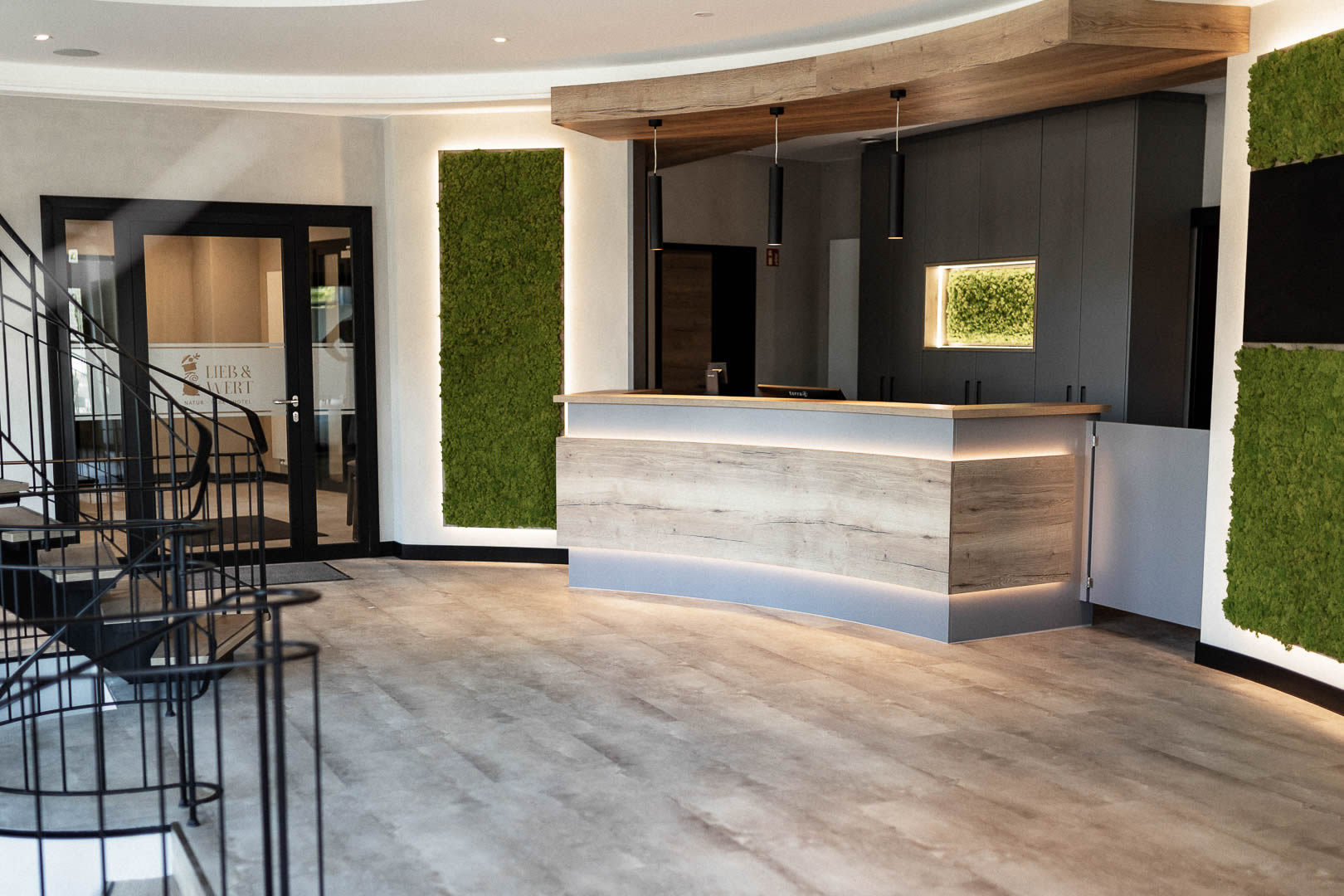Modern gestalteter Empfangsbereich mit sauber ausgeführten Maler- und Bodenlegearbeiten. Zu sehen sind helle Wand- und Deckenflächen, ein großformatiger Designboden in Steinoptik sowie eine Rezeption mit Holzoberflächen und integrierter Beleuchtung. Dunkle Rahmenelemente und grüne Wandflächen setzen zusätzliche Akzente im Innenraum.