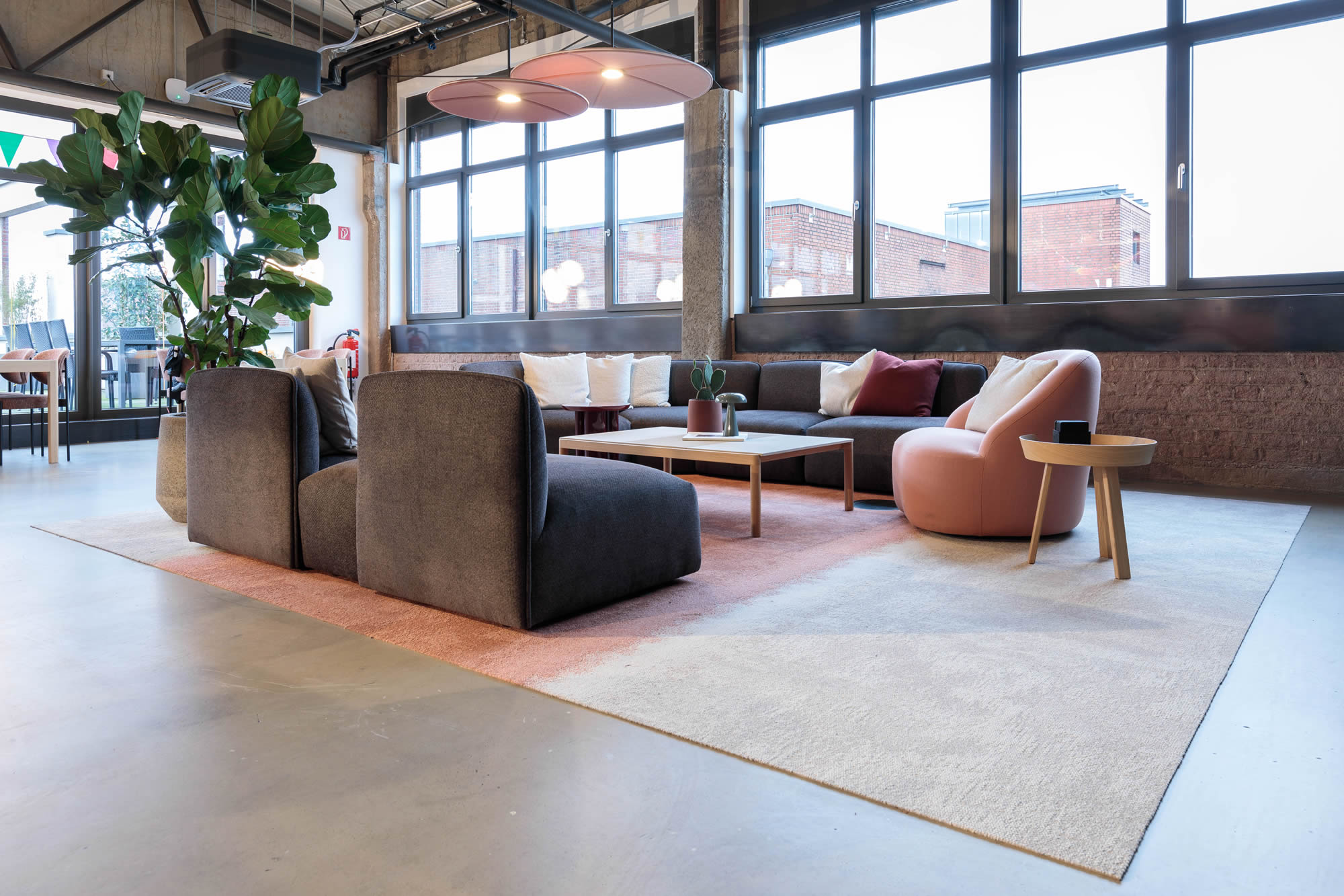 Modern gestalteter Loungebereich mit hochwertig ausgeführten Bodenlegearbeiten. Zu sehen sind eine großflächige Teppichzone in hellen und roséfarbenen Tönen, eine Sitzgruppe mit Sesseln und Sofa sowie große Fensterfronten mit viel Tageslicht. Die Aufnahme betont die saubere Ausführung der Bodenflächen und die wohnliche Gestaltung des Aufenthaltsbereichs.