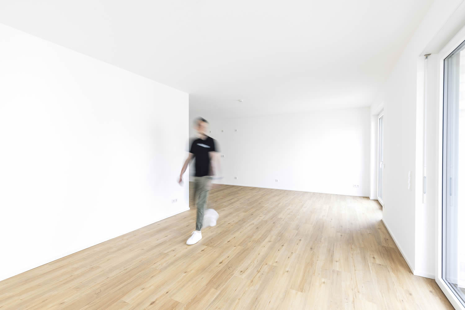 Großzügiger Wohnraum mit sauber ausgeführten Maler- und Bodenlegearbeiten. Zu sehen sind gleichmäßig weiß beschichtete Wand- und Deckenflächen, ein heller Boden in Holzoptik sowie große Fensterflächen mit viel Tageslicht. Eine unscharf dargestellte Person im Raum betont die Weite und moderne Wohnatmosphäre.