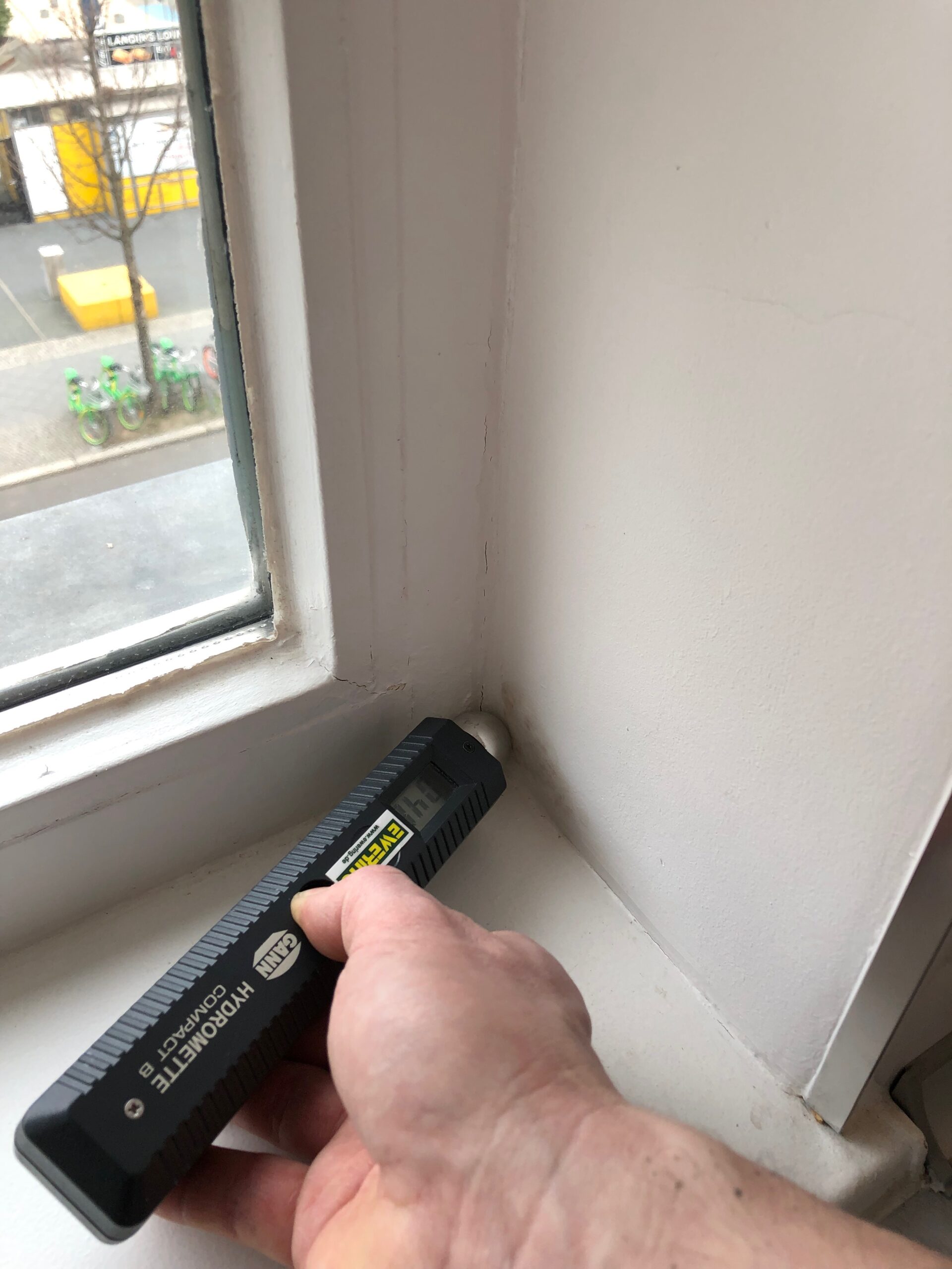 Hand misst mit einem Feuchtigkeitsmessgerät die Restfeuchte am inneren Fensterrahmen und an der angrenzenden Wand.