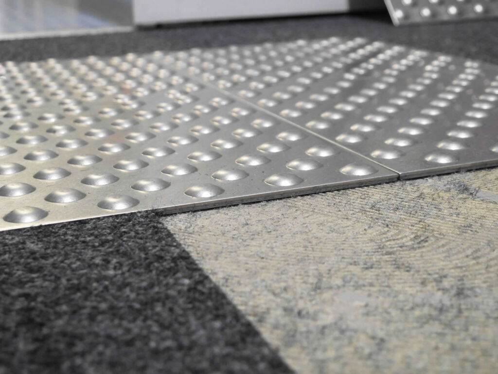 Nahaufnahme eines flächenbündig eingebauten Blindenleitsystems aus Edelstahl an einem Bodenübergang.