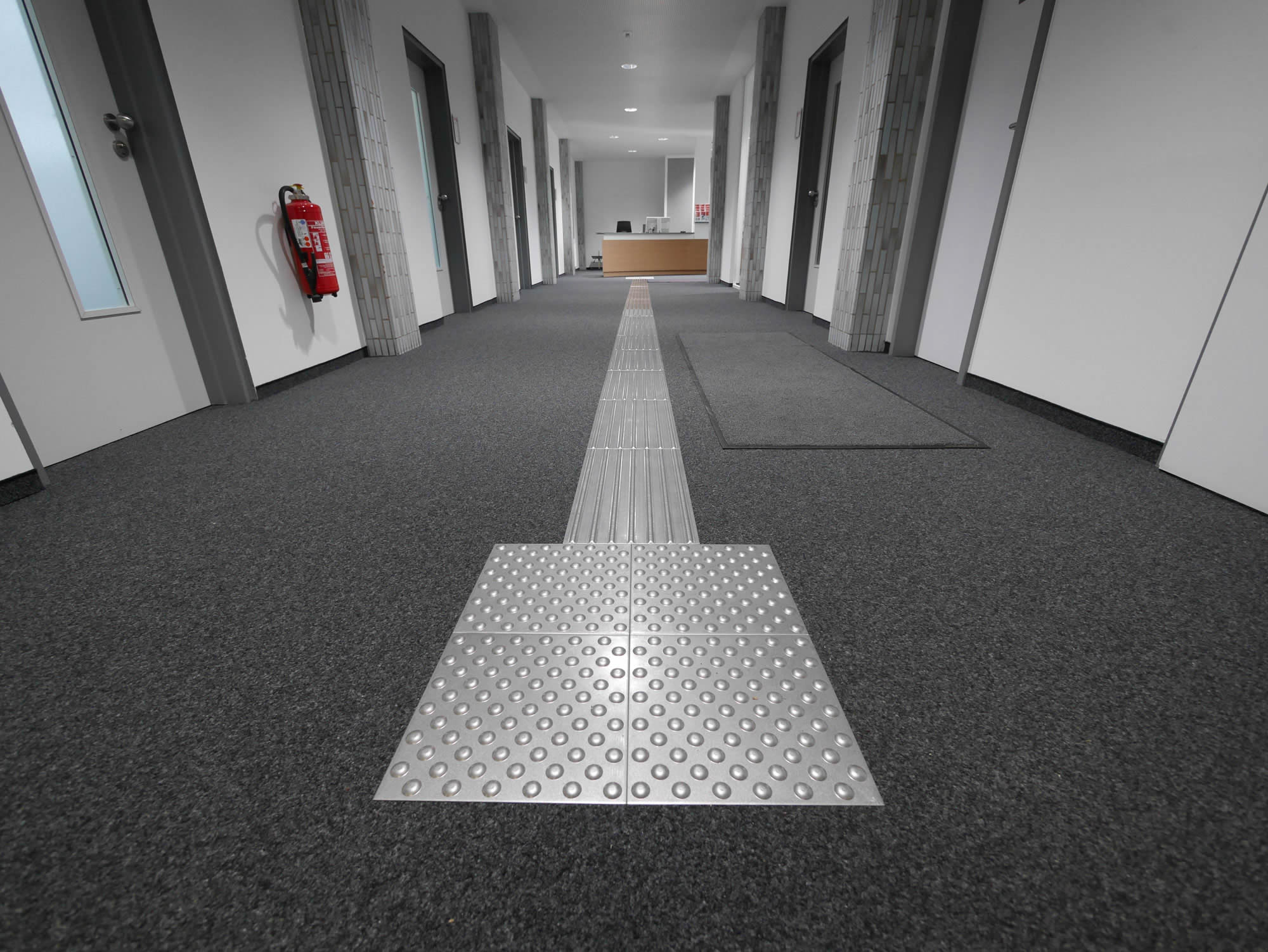 Flächenbündiges Blindenleitsystem mit Rippen- und Noppenelementen in einem Flur mit Teppichboden.