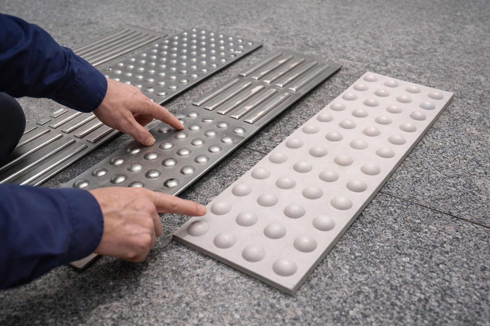 Hände zeigen auf verschiedene Rippen- und Noppenplatten für ein taktiles Blindenleitsystem.